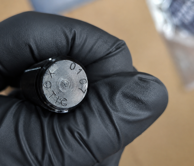 A close-up of a hand wearing a black glove holding a small cylindrical metal component with engraved markings, photographed against a blurred tan background.